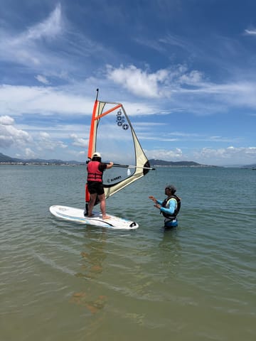 Instructor guiando a un alumno en la tabla de windsurf, en aguas poco profundas de Ponta das Canas