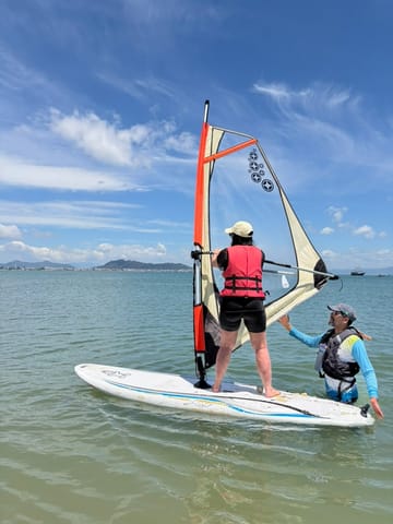 Alumno de pie sobre la tabla de windsurf con el instructor a su lado, en Ponta das Canas