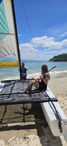 Alumna sentada en la red del catamarán Hobie Cat con la playa de Ponta das Canas de fondo