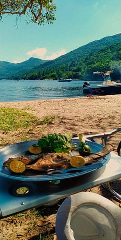 Prato de comida servido sobre mesa na praia, com a lagoa e morros ao fundo