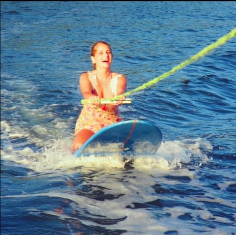 Aluna sorrindo em pé na prancha, puxada por corda atrás do barco
