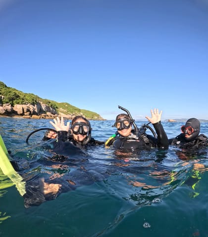 Grupo na superfície do mar com máscara e snorkel, acenando perto de uma costa rochosa