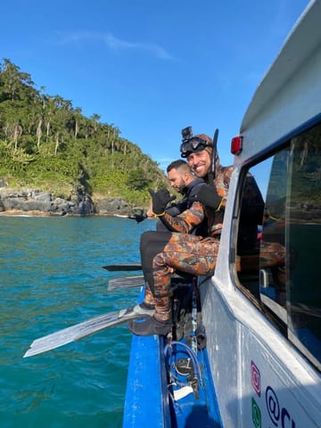 Mergulhador de roupa de borracha e nadadeiras na lateral do barco, com mata ao fundo